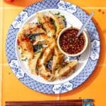 Pan-fried gyoza dumplings on a blue and white plate served with a small bowl of gyoza dipping sauce topped with sesame seeds and chilli flakes.