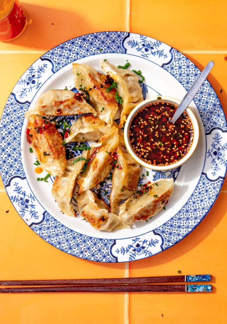 Pan-fried gyoza dumplings on a blue and white plate served with a small bowl of gyoza dipping sauce topped with sesame seeds and chilli flakes.