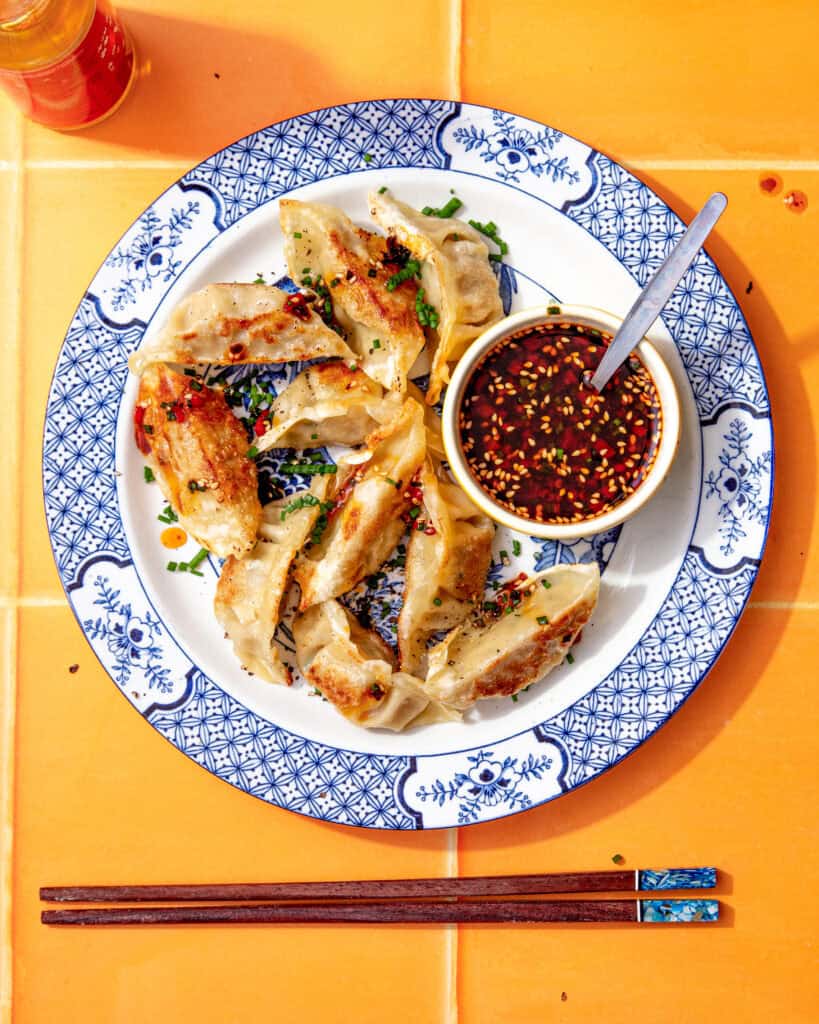 Pan-fried gyoza dumplings on a blue and white plate served with a small bowl of gyoza dipping sauce topped with sesame seeds and chilli flakes.