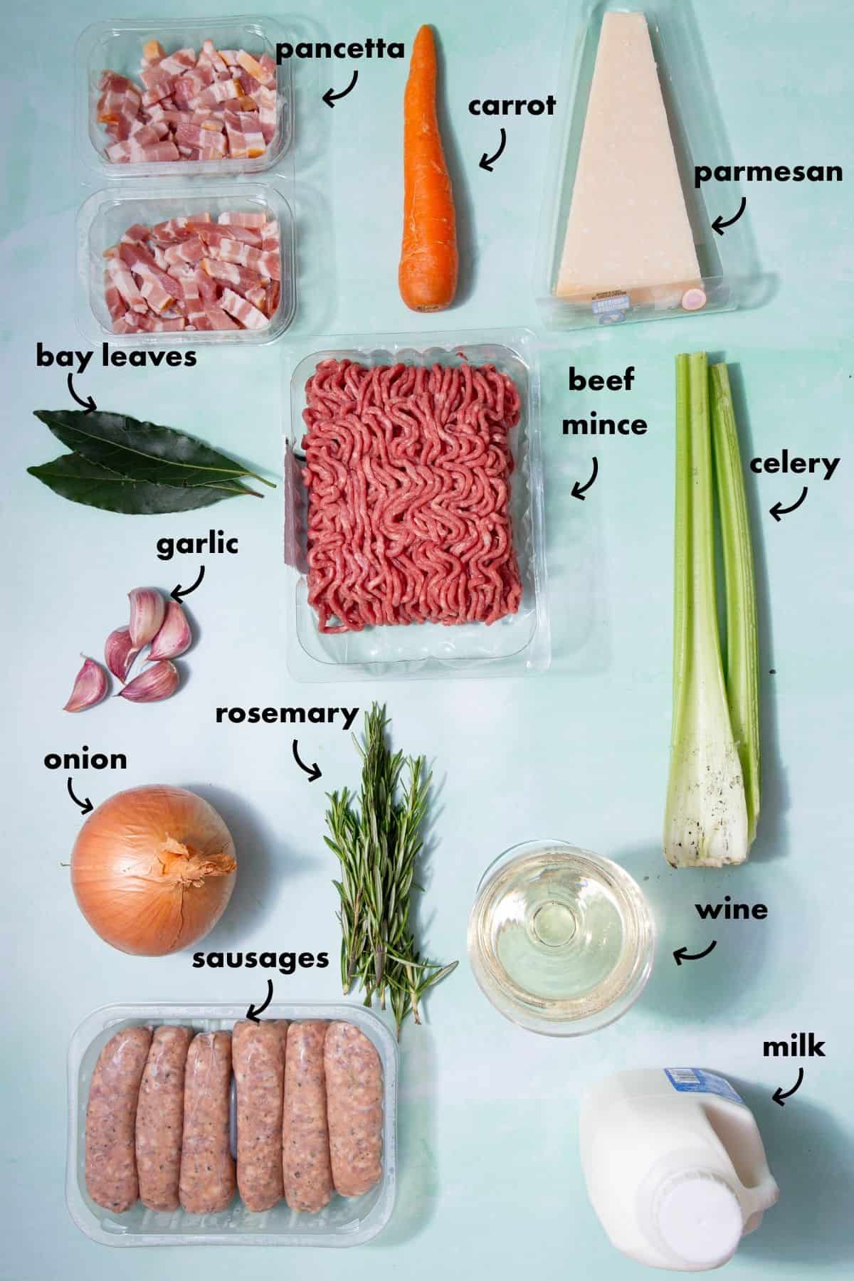 Ragu bianco ingredients laid out on a light surface including beef mince, sausages, pancetta, onion, garlic, carrot, celery, rosemary, bay leaves, parmesan, milk and white wine.