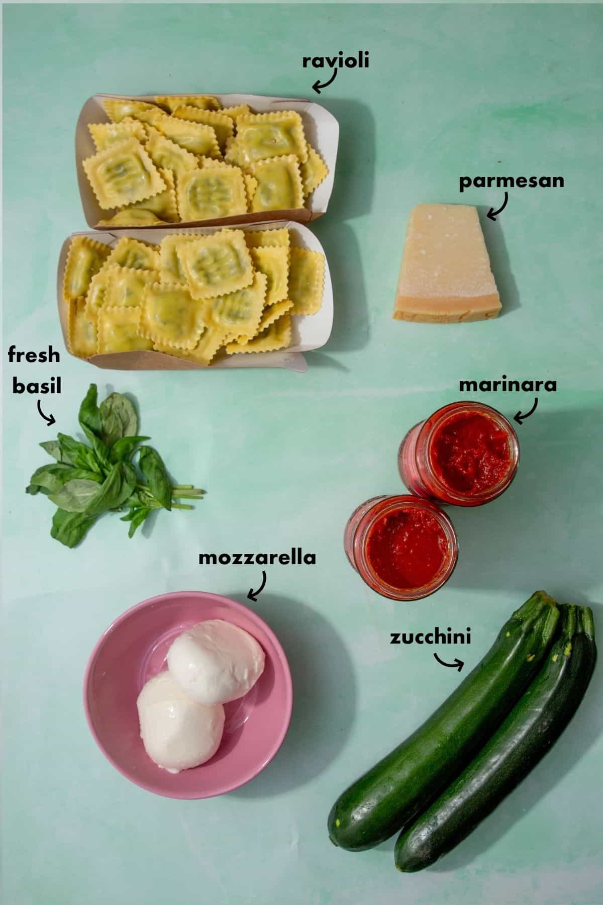 Flat lay of ingredients for baked ravioli casserole, including spinach and ricotta ravioli, mozzarella, parmesan, marinara sauce, zucchini, and fresh basil.