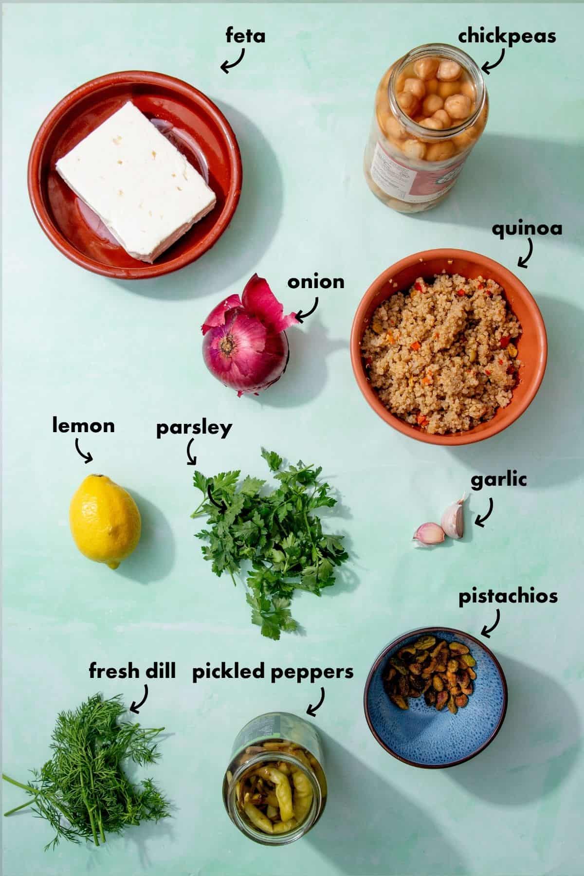 A flat lay of ingredients for the Jennifer Aniston salad on a light green surface: a block of feta, a jar of chickpeas, cooked quinoa, red onion, lemon, parsley, garlic cloves, fresh dill, pickled peppers, and pistachios. Labels identify each item.