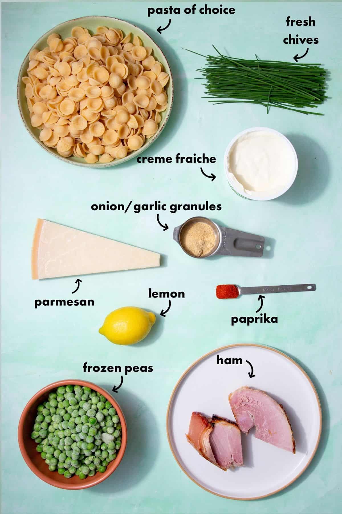 Ingredients laid out for creamy ham pasta including orecchiette, crème fraîche, peas, ham, lemon, Parmesan, paprika, garlic and onion granules, and fresh chives.