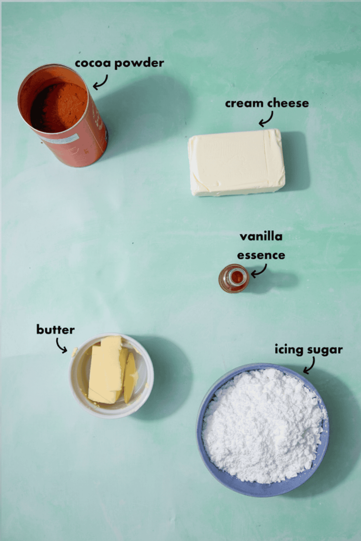 Five baking ingredients for chocolate cream cheese frosting are labeled on a light green surface: an open tin of cocoa powder, a block of cream cheese, a small bottle of vanilla essence, a bowl of butter, and a bowl of icing sugar.