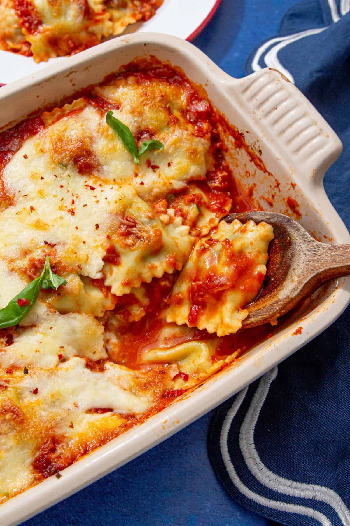 Close-up of baked ravioli casserole in a cream dish, with golden melted cheese, marinara sauce, and fresh basil leaves.