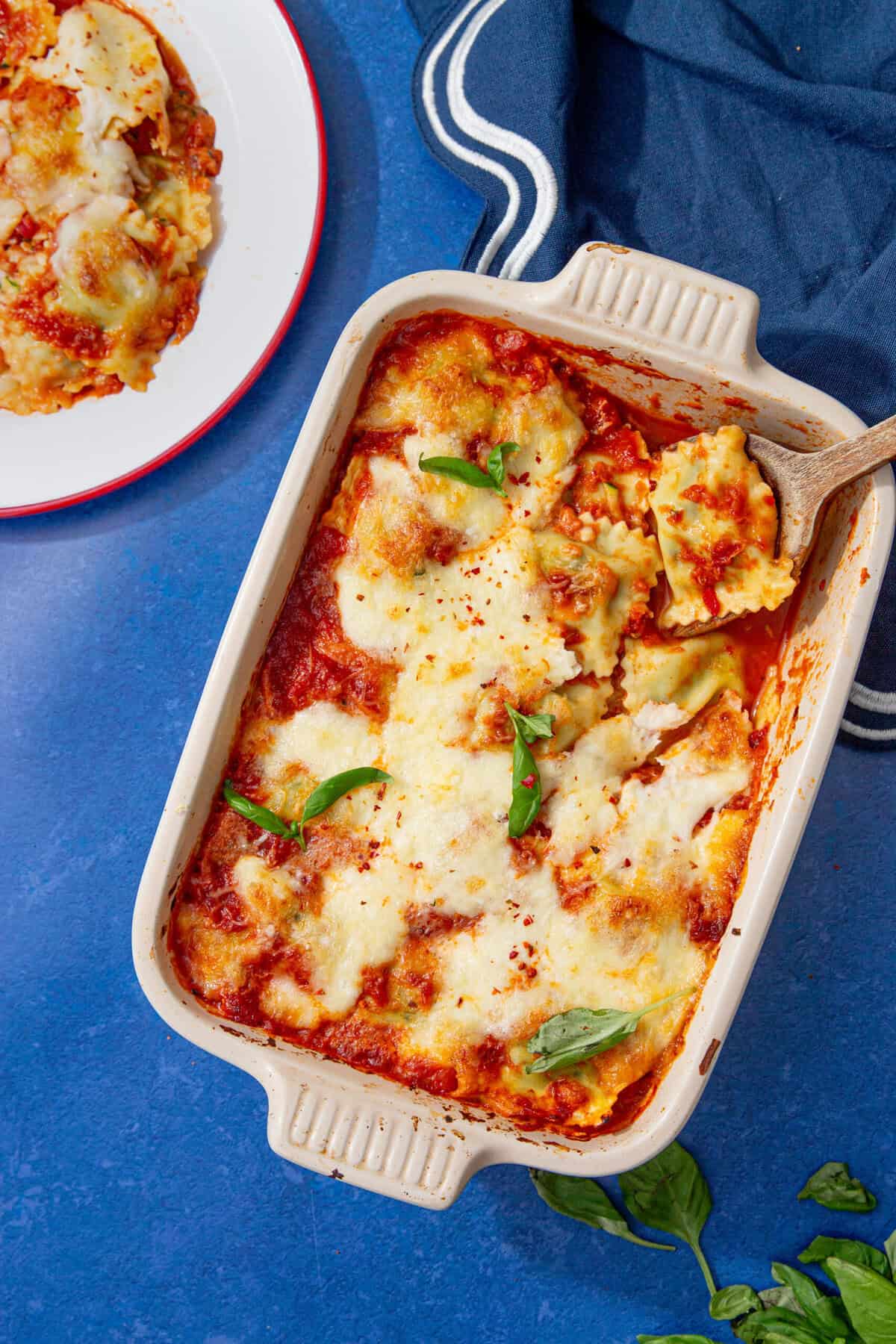 Overhead view of a baked ravioli casserole topped with melted mozzarella and basil, served next to a plate of ravioli on a blue tablecloth.