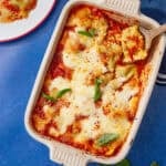 Overhead view of baked ravioli casserole in a cream dish topped with melted mozzarella, marinara, and fresh basil, with a serving of ravioli on a plate beside it on a blue surface.