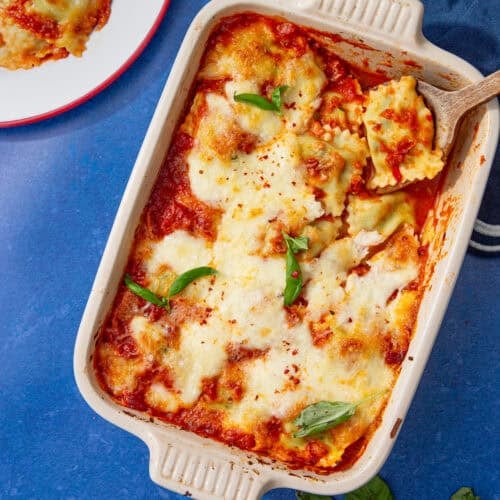 Overhead view of baked ravioli casserole in a cream dish topped with melted mozzarella, marinara, and fresh basil, with a serving of ravioli on a plate beside it on a blue surface.