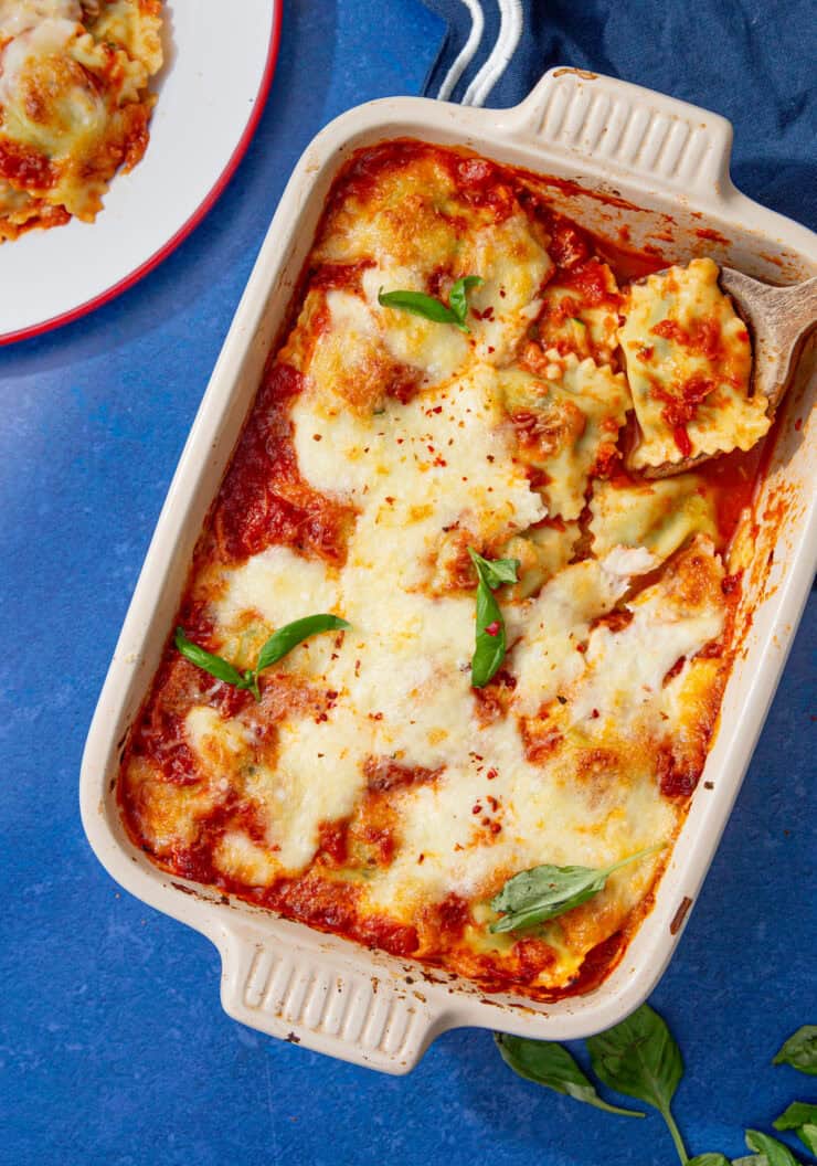 Overhead view of baked ravioli casserole in a cream dish topped with melted mozzarella, marinara, and fresh basil, with a serving of ravioli on a plate beside it on a blue surface.