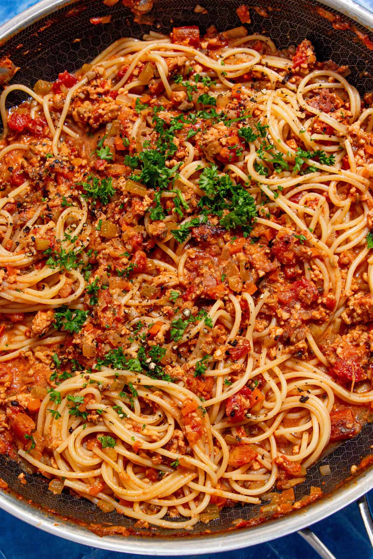 A close-up of cooked spaghetti topped with a chunky tomato and ground meat sauce, garnished with chopped parsley, served in a large skillet.
