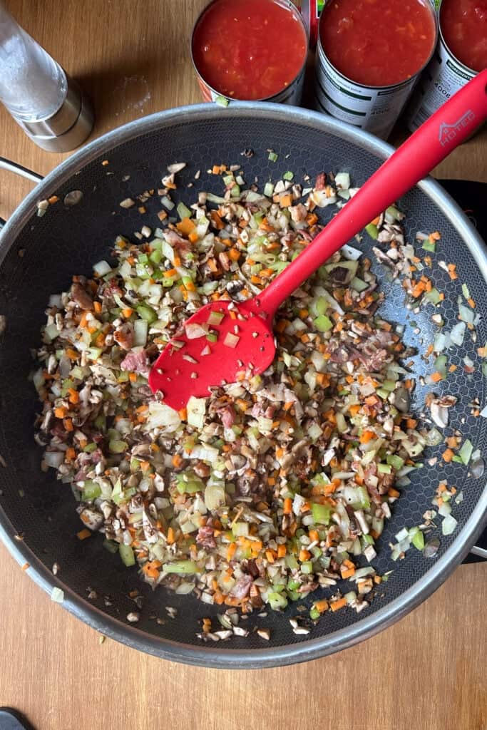 A frying pan with chopped onions, carrots, celery, mushrooms, and bacon being sautéed, stirred with a red spatula. Open cans of tomatoes are visible on the wooden countertop.