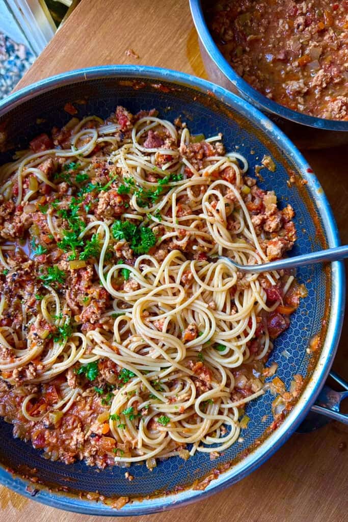 A pan of spaghetti mixed with meat sauce garnished with chopped parsley, sits on a wooden table beside a pot filled with extra sauce. A metal fork rests in the pan.