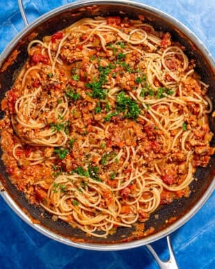 A skillet filled with cooked spaghetti mixed with a chunky tomato and ground meat sauce, garnished with chopped parsley, sits on a bright blue tiled surface.