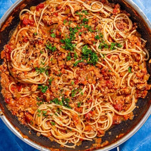 A skillet filled with cooked spaghetti mixed with a chunky tomato and ground meat sauce, garnished with chopped parsley, sits on a bright blue tiled surface.