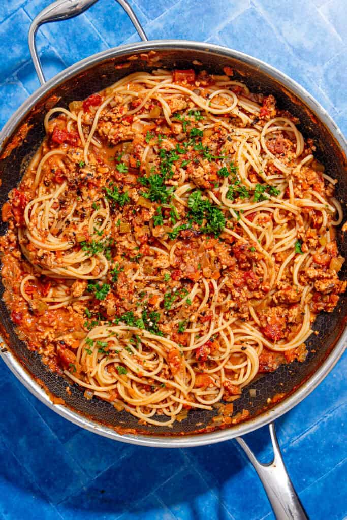 A skillet filled with cooked spaghetti mixed with a chunky tomato and ground meat sauce, garnished with chopped parsley, sits on a bright blue tiled surface.