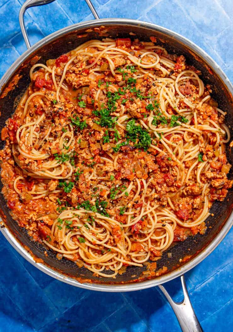 A skillet filled with cooked spaghetti mixed with a chunky tomato and ground meat sauce, garnished with chopped parsley, sits on a bright blue tiled surface.