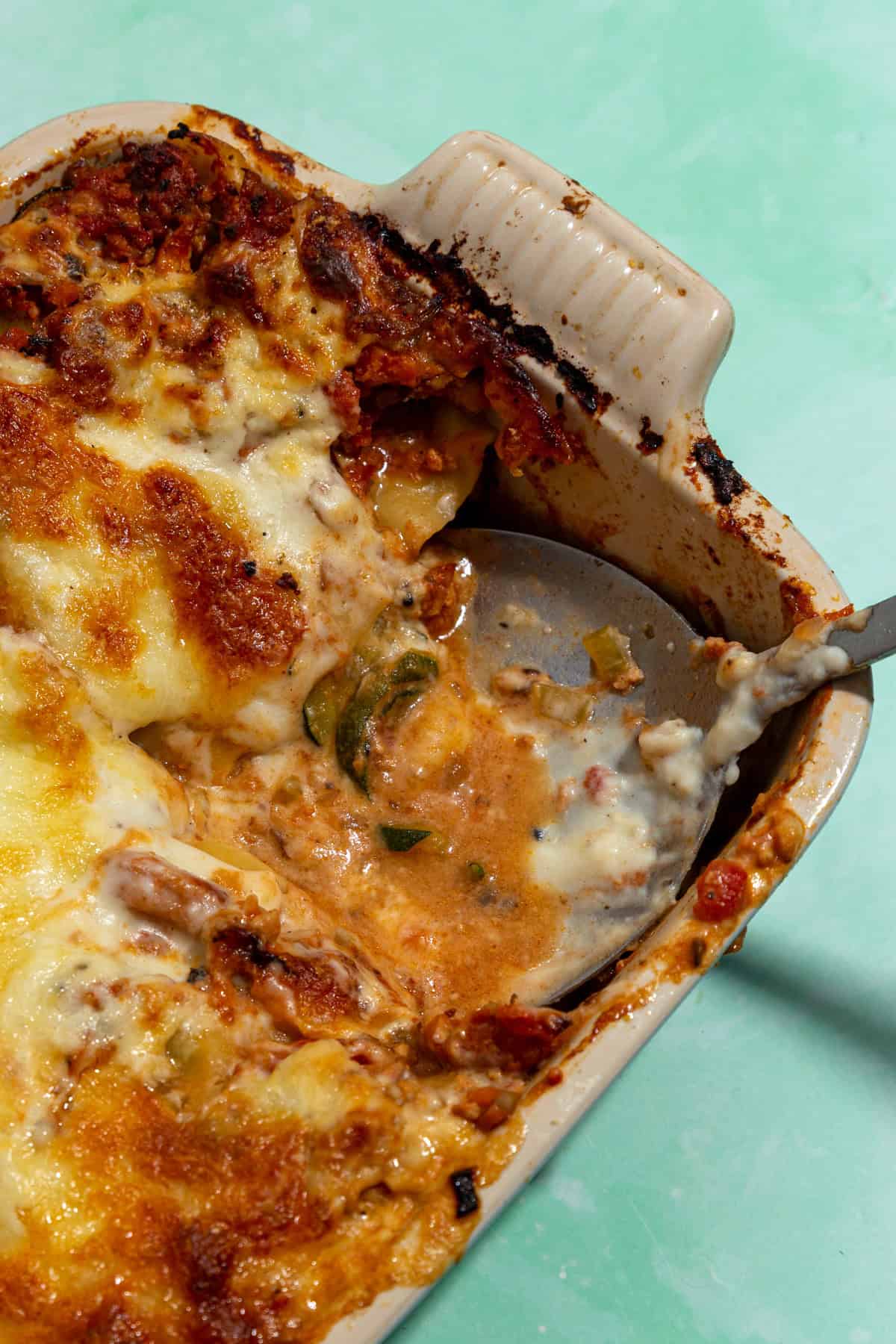 A close-up of a cheesy baked lasagna in a ceramic dish, with roasted corn blended into the layers. A serving spoon scoops out a portion, revealing sauce, cheese, pasta, and corn on a mint green surface.