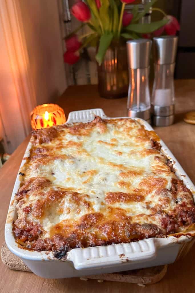 A freshly baked lasagna with a golden, melted cheese topping and hints of roasted corn sits in a white ceramic dish on a wooden table, accompanied by a lit candle, salt and pepper shakers, and a vase of red tulips in the background.