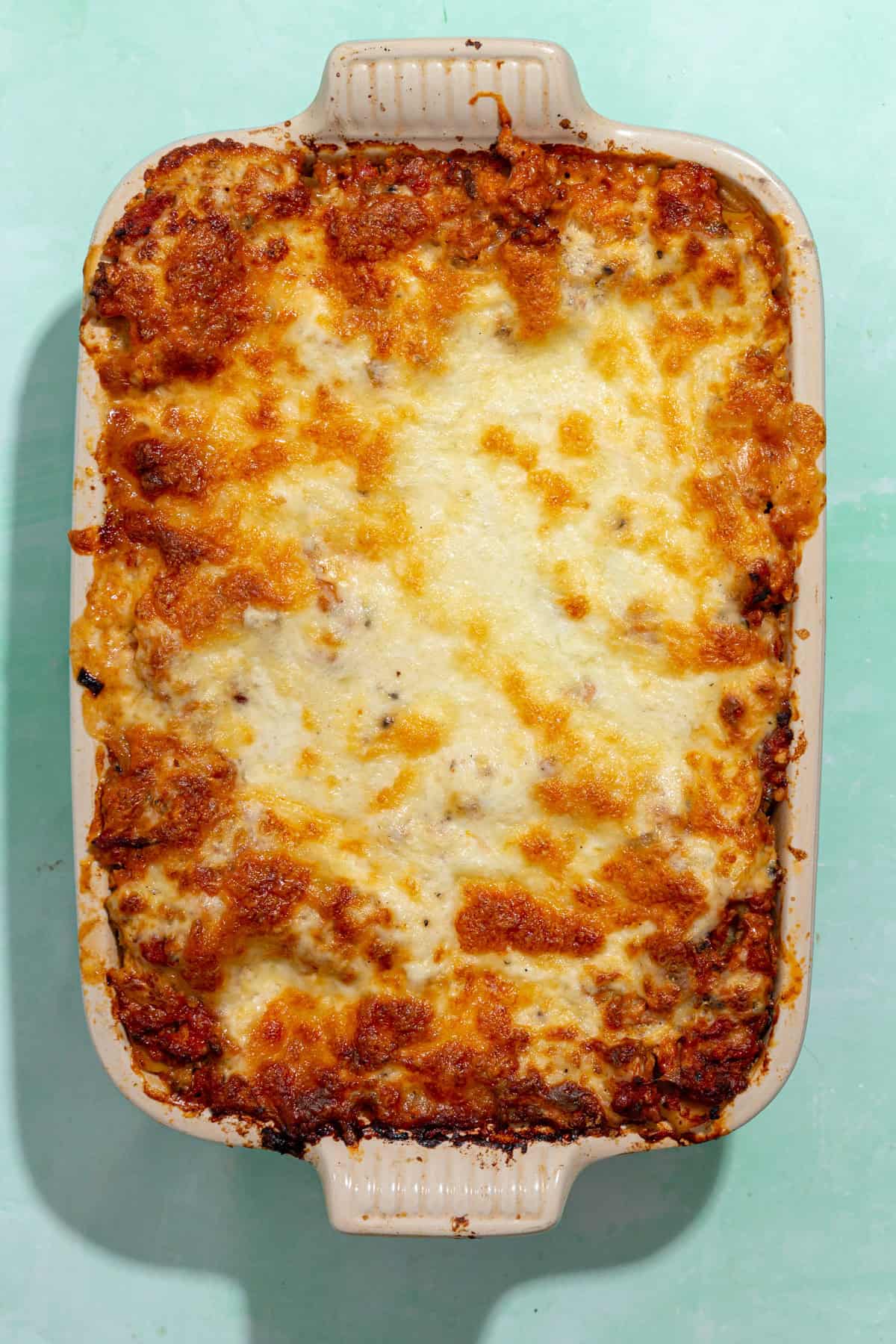 A rectangular baking dish filled with golden-brown, cheesy baked lasagna, featuring roasted corn and a bubbly, slightly browned top layer, sits on a light green surface.