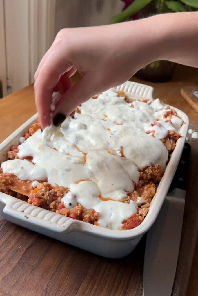 A hand sprinkles cheese over a casserole dish filled with lasagna topped with white sauce and roasted corn, sitting on a wooden table.