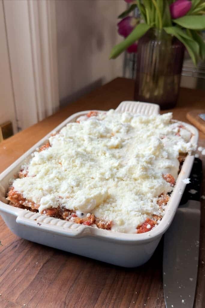 A rectangular baking dish filled with lasagna topped with a thick layer of grated cheese and roasted corn sits on a wooden surface. In the background, there is a glass vase with purple tulips.