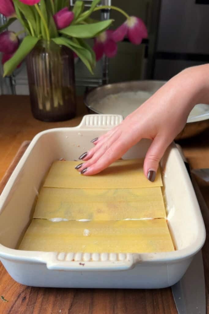 A hand places uncooked lasagna noodles into a white baking dish on a wooden surface, ready to layer with roasted corn. A vase with pink tulips and kitchen items are visible in the background.