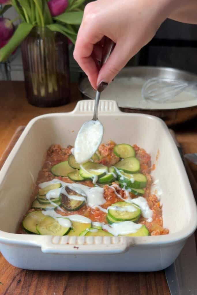 A hand holds a spoon, drizzling white sauce over sliced zucchini, tomato mixture, and roasted corn in a rectangular baking dish on a wooden counter. A vase with flowers and kitchen utensils are visible in the background.