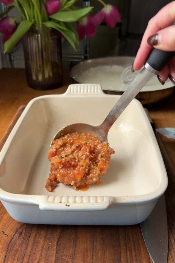 A hand holds a ladle of tomato-based meat sauce over a white ceramic baking dish, ready to spread. In the background, Roasted Corn sits near a glass vase with pink flowers on a wooden table.
