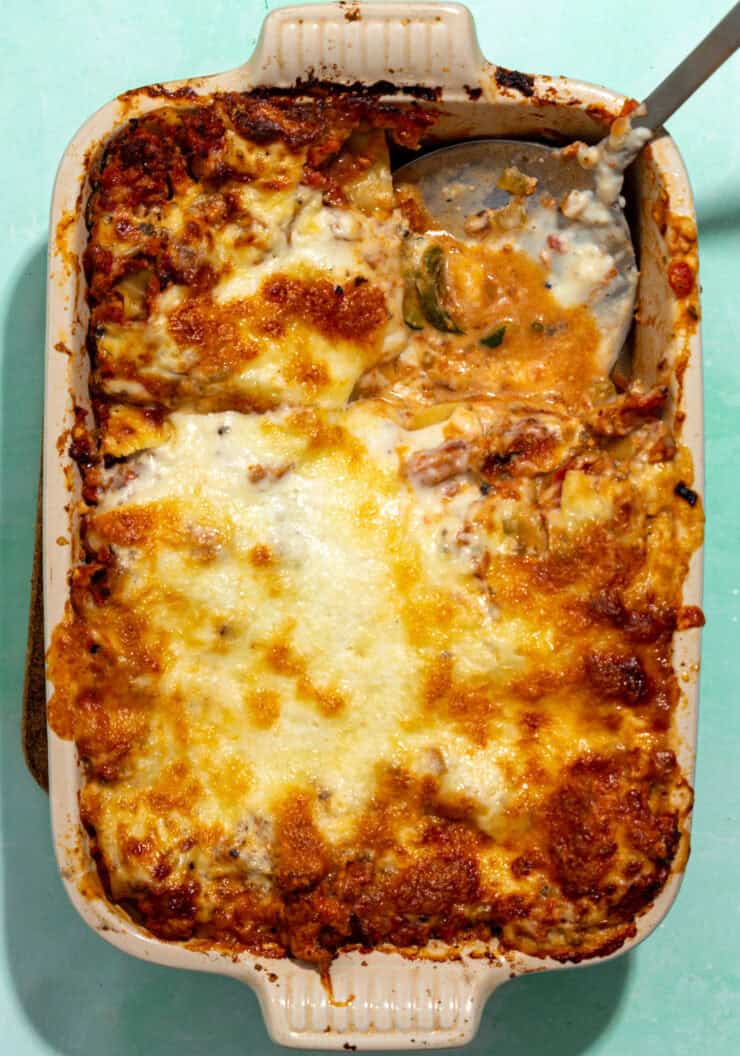A casserole dish filled with baked lasagna, topped with melted, golden-brown cheese. A serving has been scooped out, revealing layers of pasta, sauce, cheese, and sweet roasted corn nestled between.