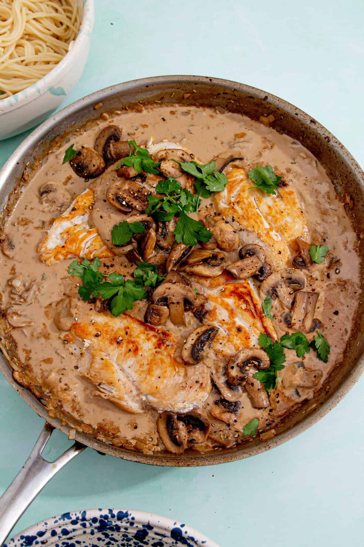 A skillet filled with browned chicken breasts in a creamy mushroom sauce, garnished with fresh parsley—the perfect cream of mushroom chicken. A bowl of cooked spaghetti sits nearby on a light blue surface.