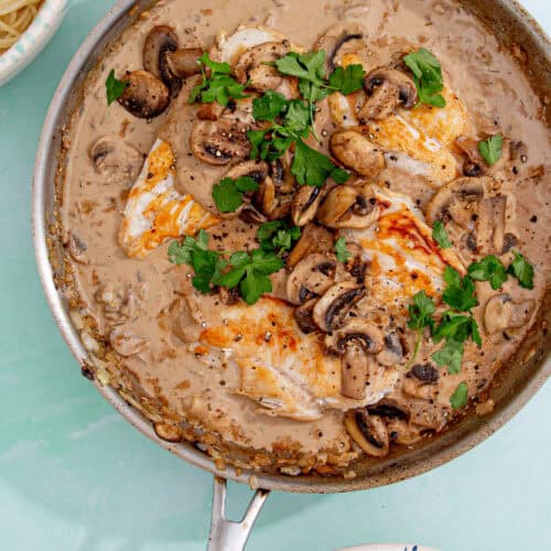 A skillet filled with cream of mushroom chicken breasts in a creamy sauce, garnished with fresh parsley. Nearby are bowls of cooked spaghetti and green peas.