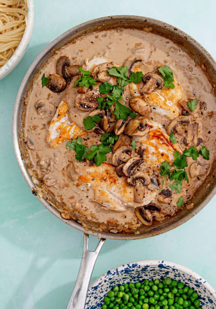 A skillet filled with cream of mushroom chicken breasts in a creamy sauce, garnished with fresh parsley. Nearby are bowls of cooked spaghetti and green peas.