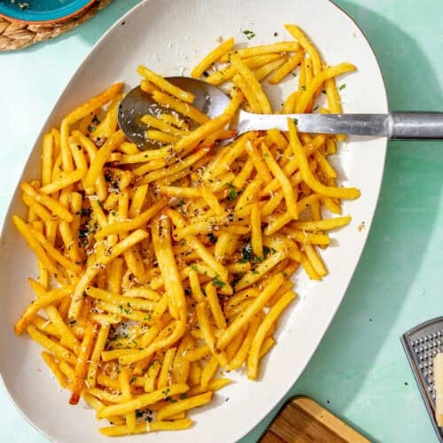 A platter of garlic fries topped with grated parmesan and chopped parsley, with a serving spoon beside them and a bowl of leftover garlic butter in the background.