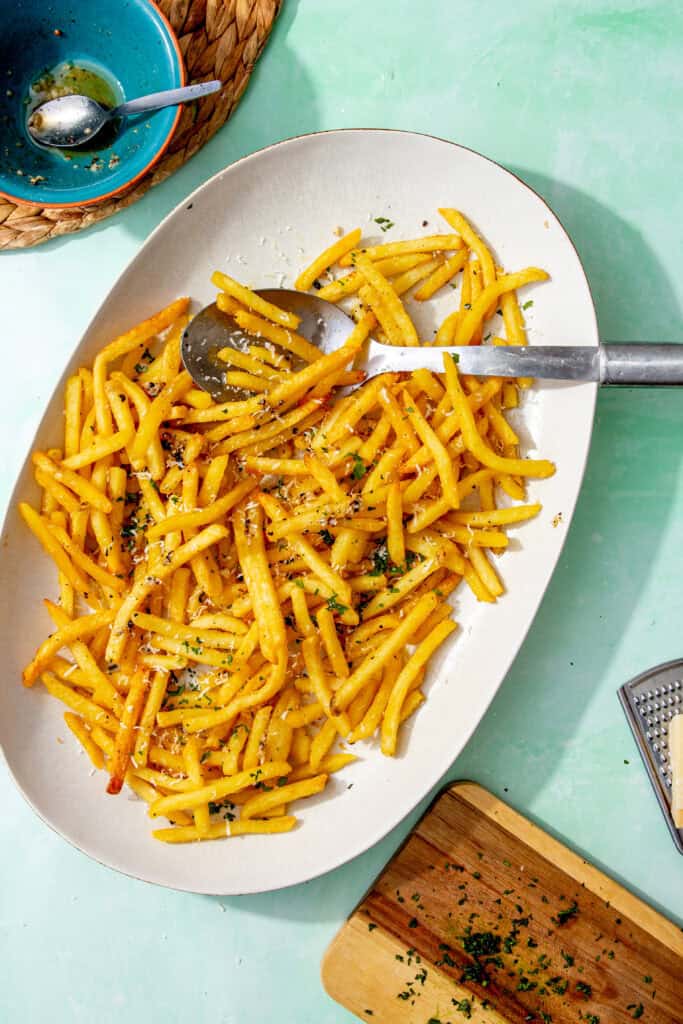 A platter of garlic fries topped with grated parmesan and chopped parsley, with a serving spoon beside them and a bowl of leftover garlic butter in the background.