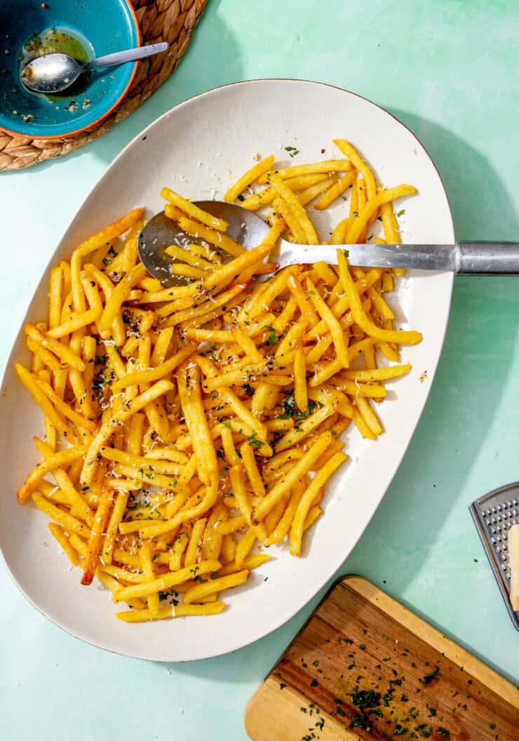 A platter of garlic fries topped with grated parmesan and chopped parsley, with a serving spoon beside them and a bowl of leftover garlic butter in the background.