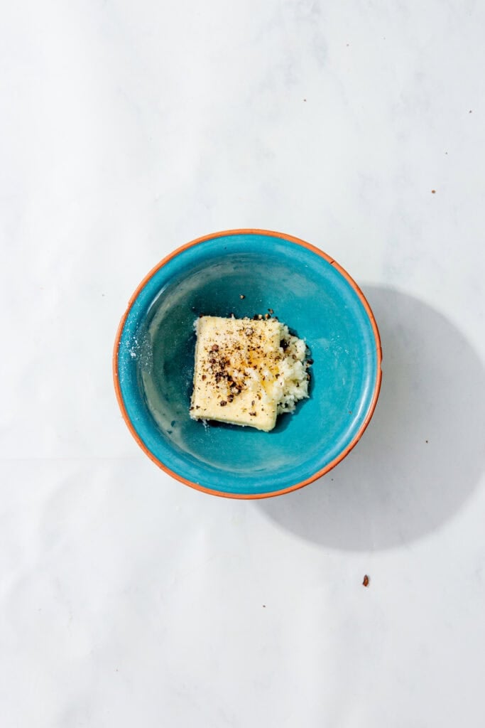 A blue bowl with butter, minced garlic, salt, and cracked black pepper before melting.