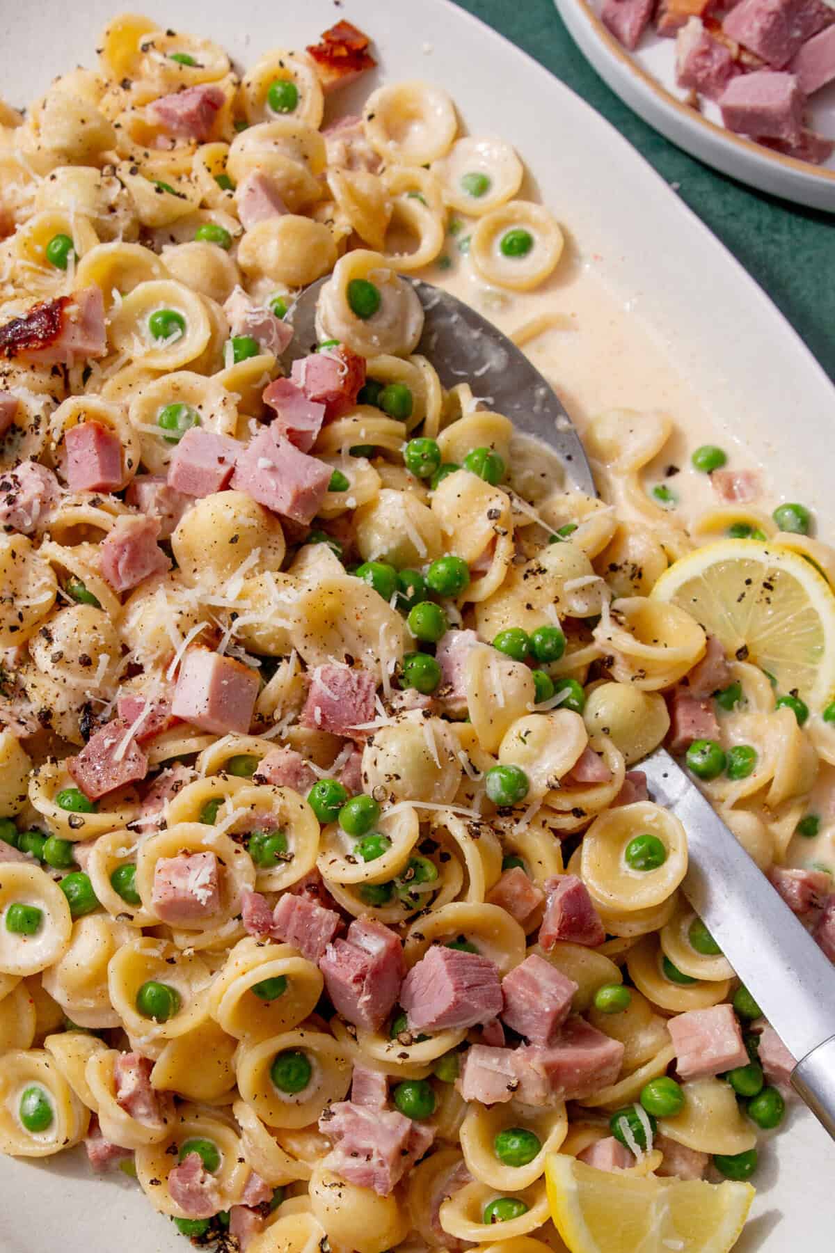 Ham pasta with peas and cheese served on a white platter, spooned ready to serve with lemon wedges on the side.