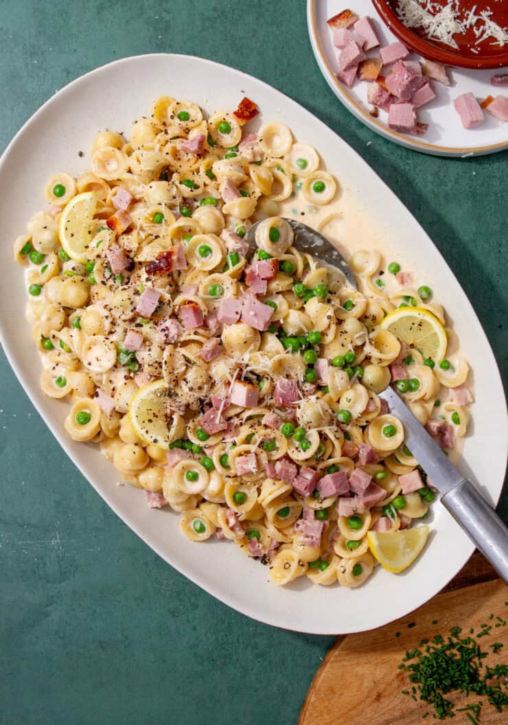 Large serving platter of creamy leftover ham pasta with peas, lemon slices, and Parmesan, ready to serve with chopped chives on the side.