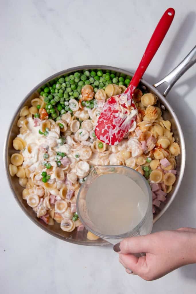 Skillet filled with cooked pasta, peas, chopped ham, crème fraîche, and spices before mixing.