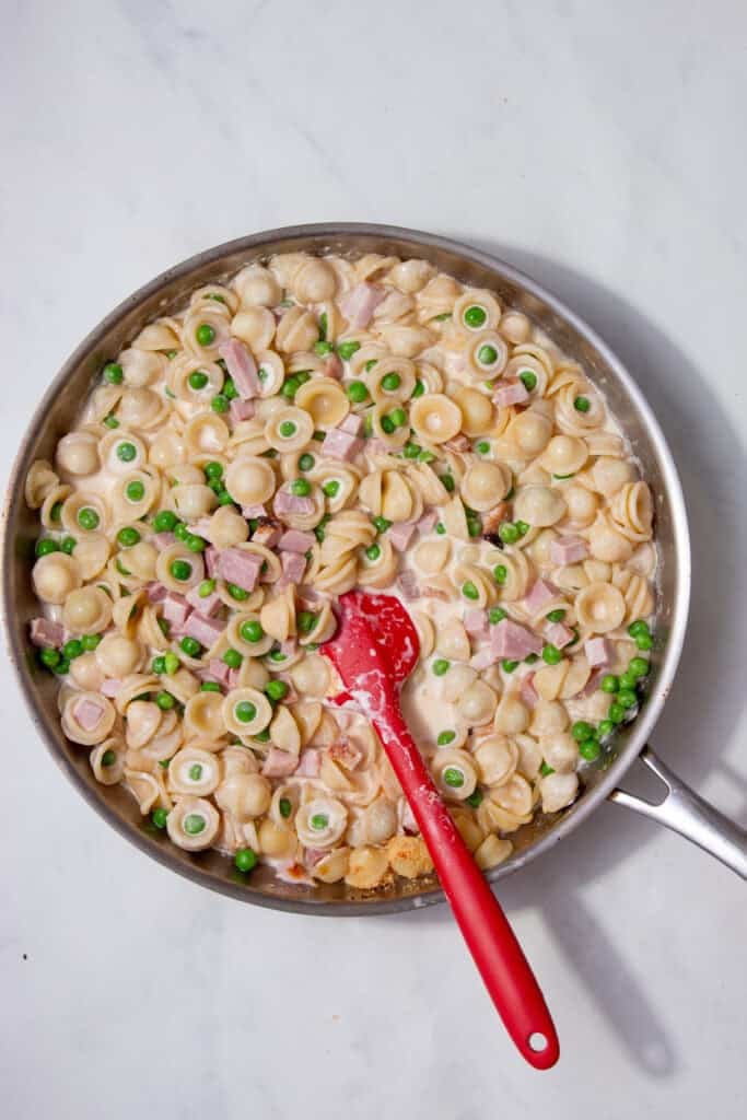Hand pouring reserved pasta water into the pan of pasta, ham, and peas.