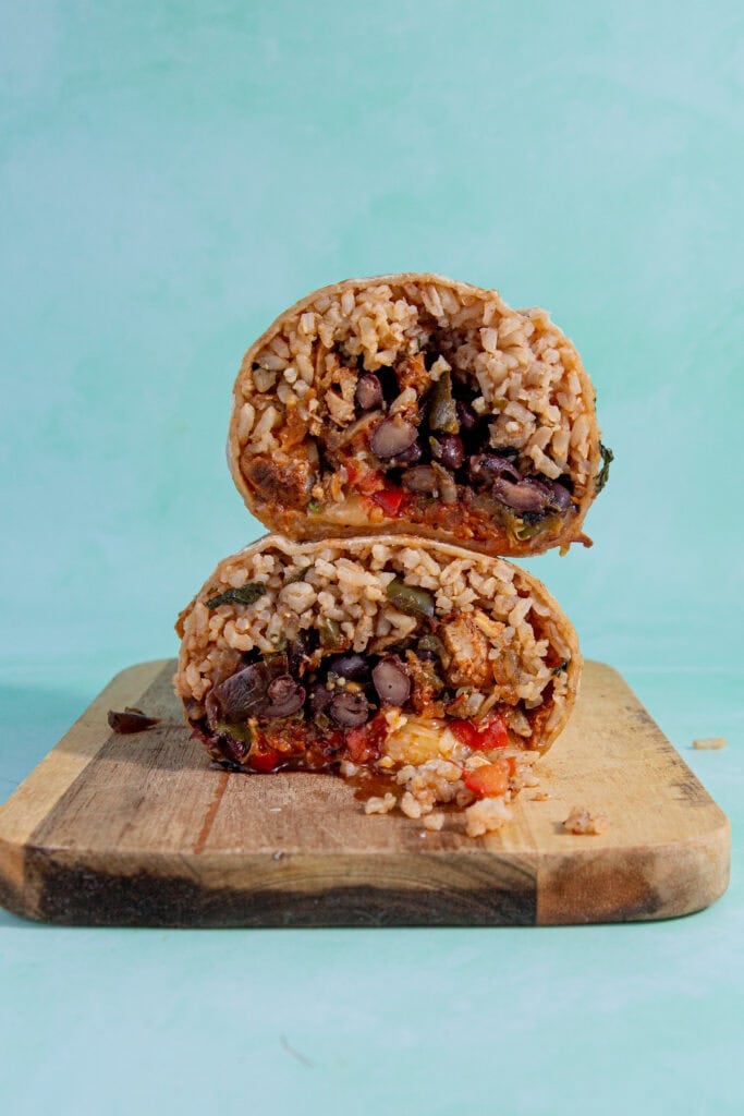 A burrito cut in half, showing rice, black beans, vegetables, and sauce inside, stacked on a wooden board against a light turquoise background—perfect for learning how to reheat a burrito and enjoy leftovers.