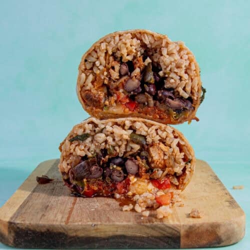 Two halves of a burrito, filled with rice, black beans, vegetables, and sauce, are stacked on a wooden cutting board against a light turquoise background—perfect for learning how to reheat a burrito while keeping it fresh and delicious.