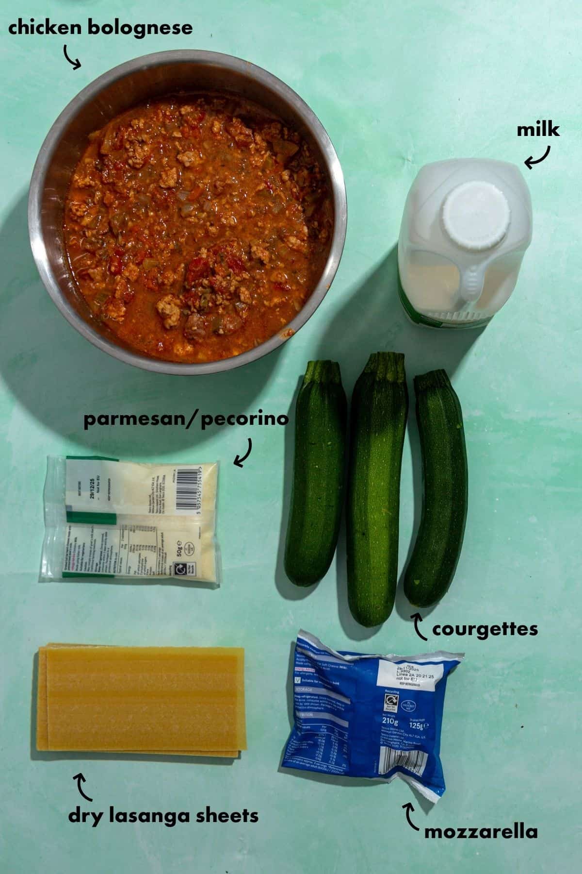 Ingredients for a lasagna are laid out on a green surface: a bowl of chicken bolognese, roasted corn, a carton of milk, three courgettes, parmesan/pecorino cheese, mozzarella, and dry lasagna sheets—all clearly labeled.