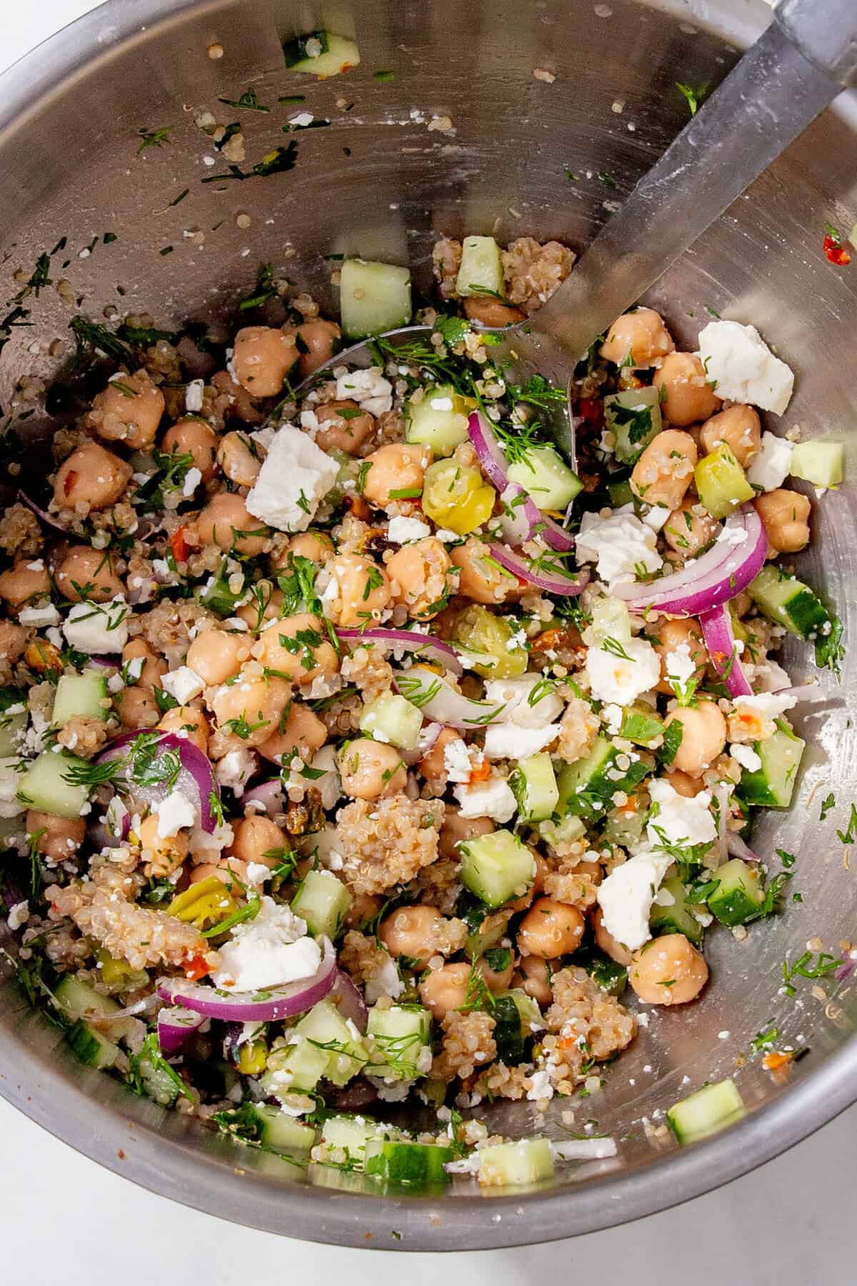 A metal bowl filled with a colorful salad of chickpeas, cucumber, red onion, feta cheese, fresh herbs, and quinoa, with a spoon for mixing.