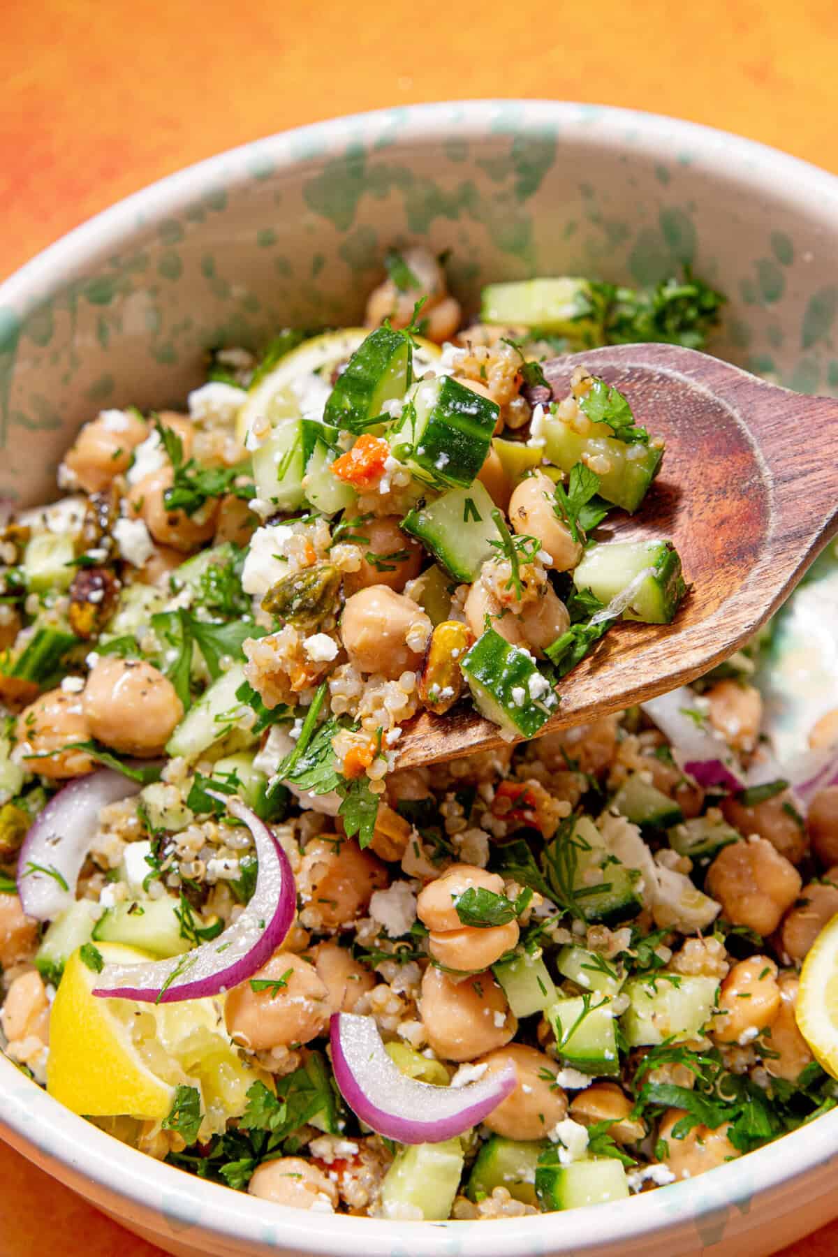 A close-up of a colorful Jennifer Aniston salad with chickpeas, diced cucumber, red onion, herbs, and feta cheese, being scooped with a wooden spoon from a bowl. A lemon wedge is visible in the salad.
