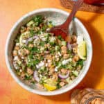 A bowl of the Jennifer Aniston salad with chickpeas, sliced red onions, cucumber, parsley, crumbled feta, and lemon wedges, with a wooden spoon resting in the bowl on an orange tabletop.