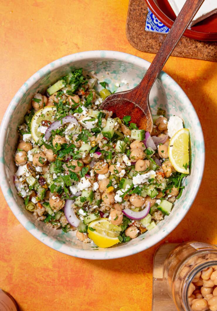A bowl of the Jennifer Aniston salad with chickpeas, sliced red onions, cucumber, parsley, crumbled feta, and lemon wedges, with a wooden spoon resting in the bowl on an orange tabletop.