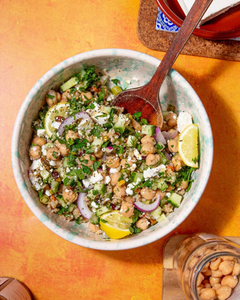 A bowl of the Jennifer Aniston salad with chickpeas, sliced red onions, cucumber, parsley, crumbled feta, and lemon wedges, with a wooden spoon resting in the bowl on an orange tabletop.