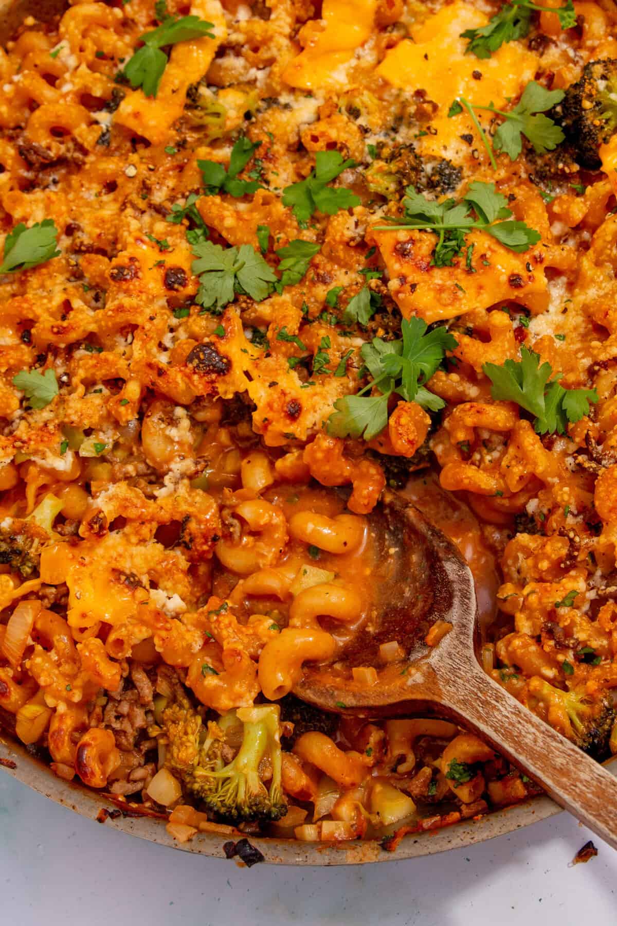 A close-up of cheesy baked mince pasta with broccoli, golden melted cheese, fresh parsley, and breadcrumbs in a pan, with a wooden spoon scooping some out.
