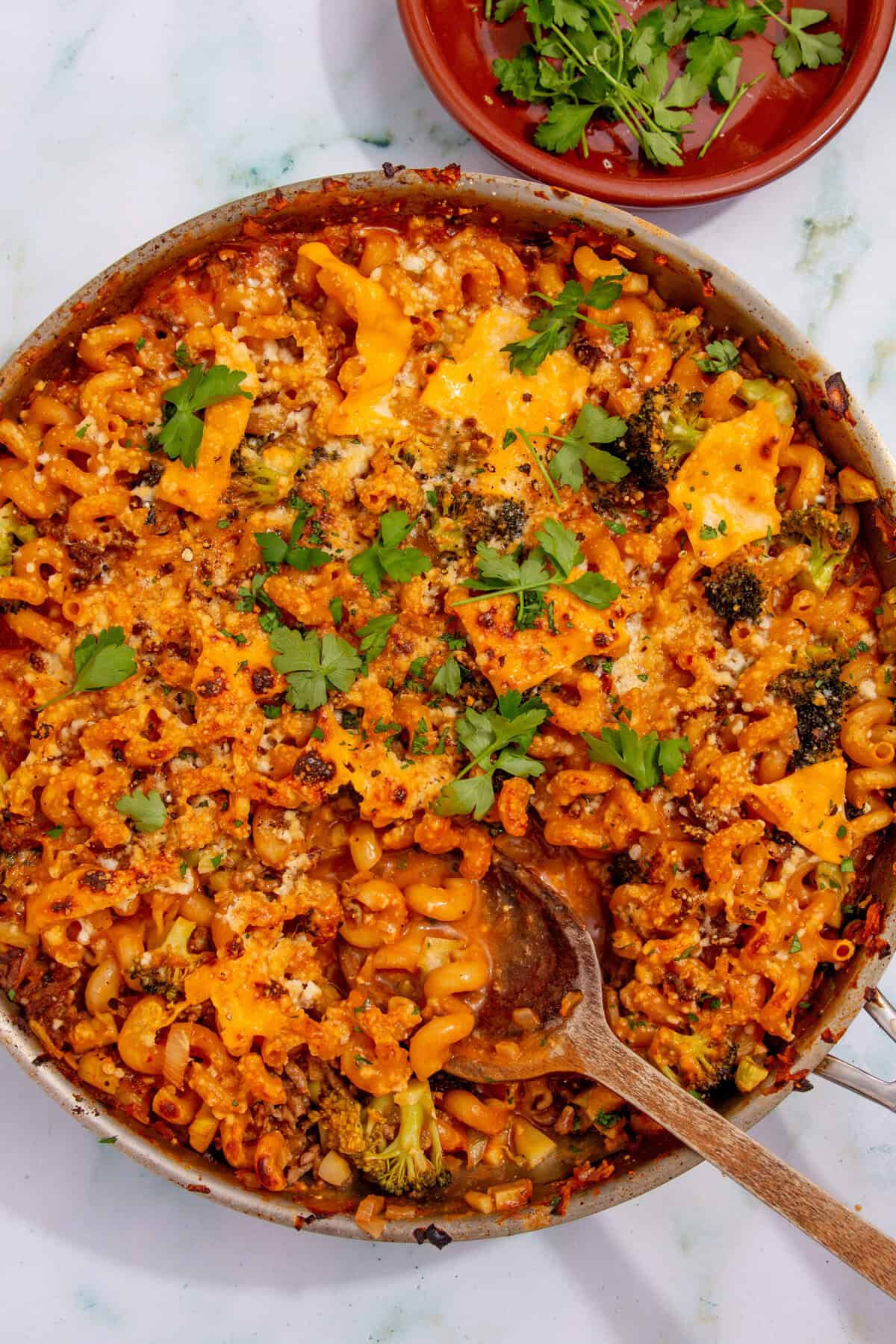 A skillet filled with mince pasta baked in tomato sauce, topped with melted cheese, broccoli, herbs, and fresh parsley. A wooden spoon rests in the pan, with a small dish of extra parsley nearby.